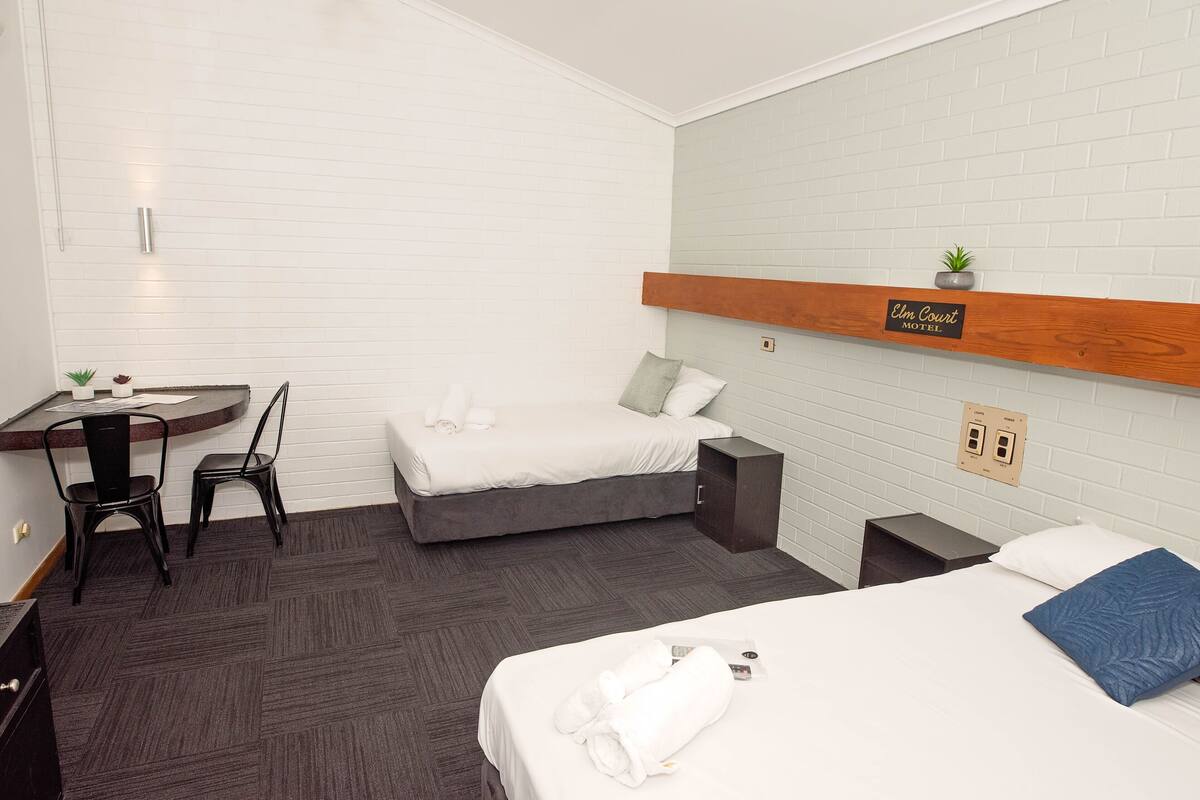 A hotel room with a bed, a desk with a chair, a small sink, and a wooden shelf with a sign.