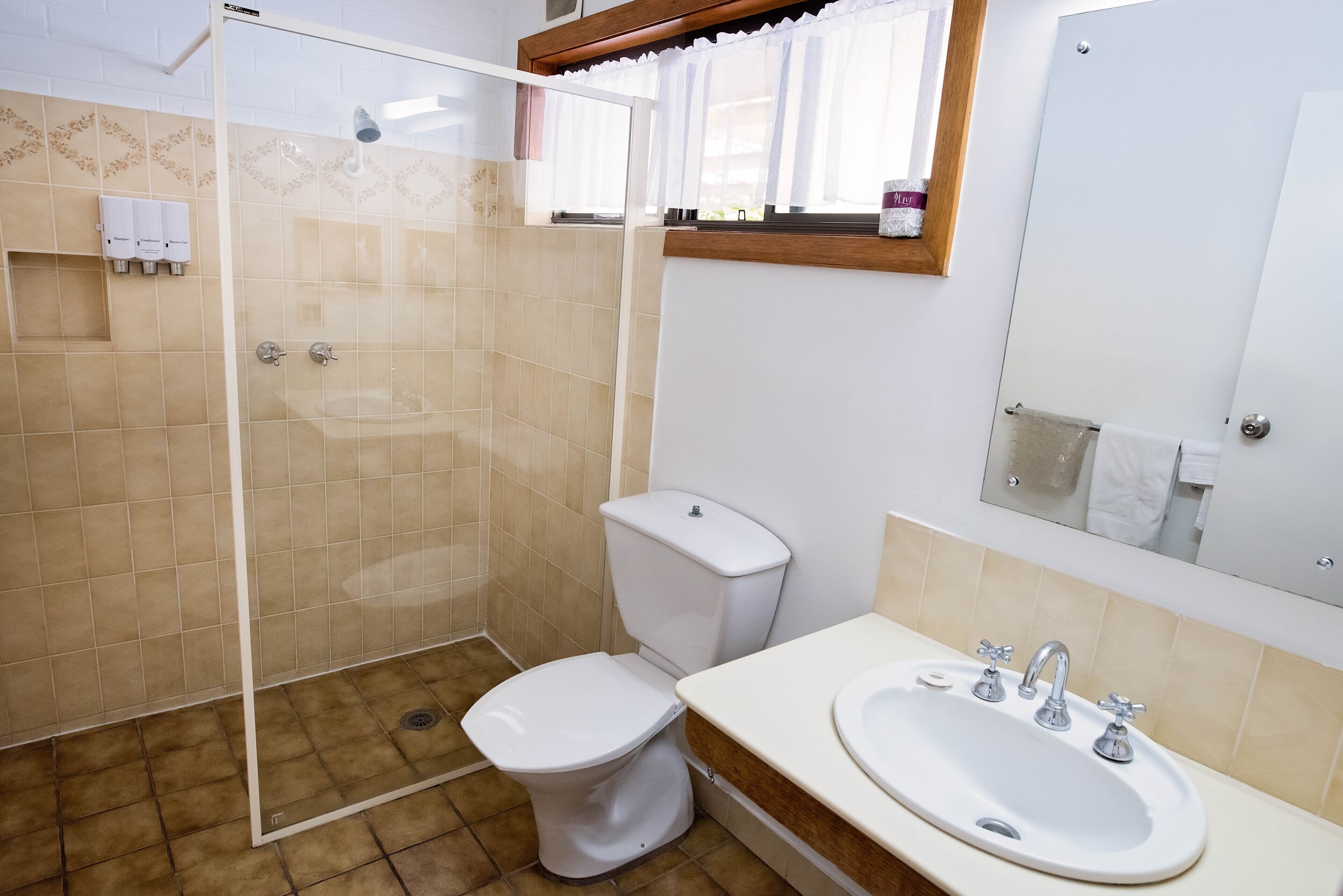 Un baño con ducha de vidrio, inodoro, lavamanos con espejo y ventana con cortinas.