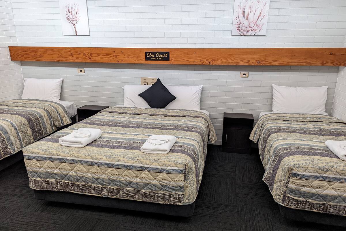 A hotel room with three beds, each with patterned bedding and white pillows. There is a wooden headboard with a sign above it.