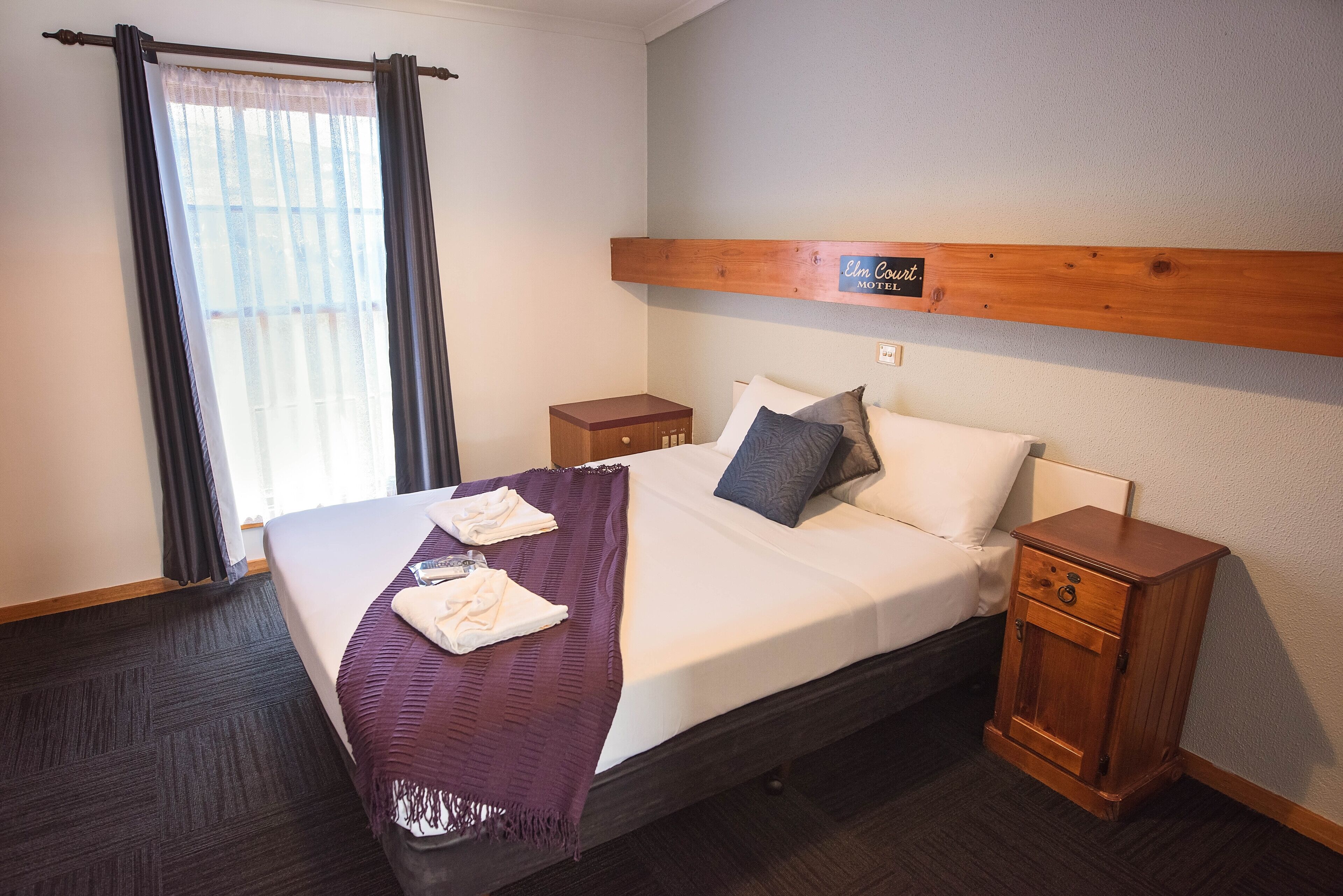 Cama bem arrumada com manta roxa, dois travesseiros e uma mesa de cabeceira de madeira com gavetas.