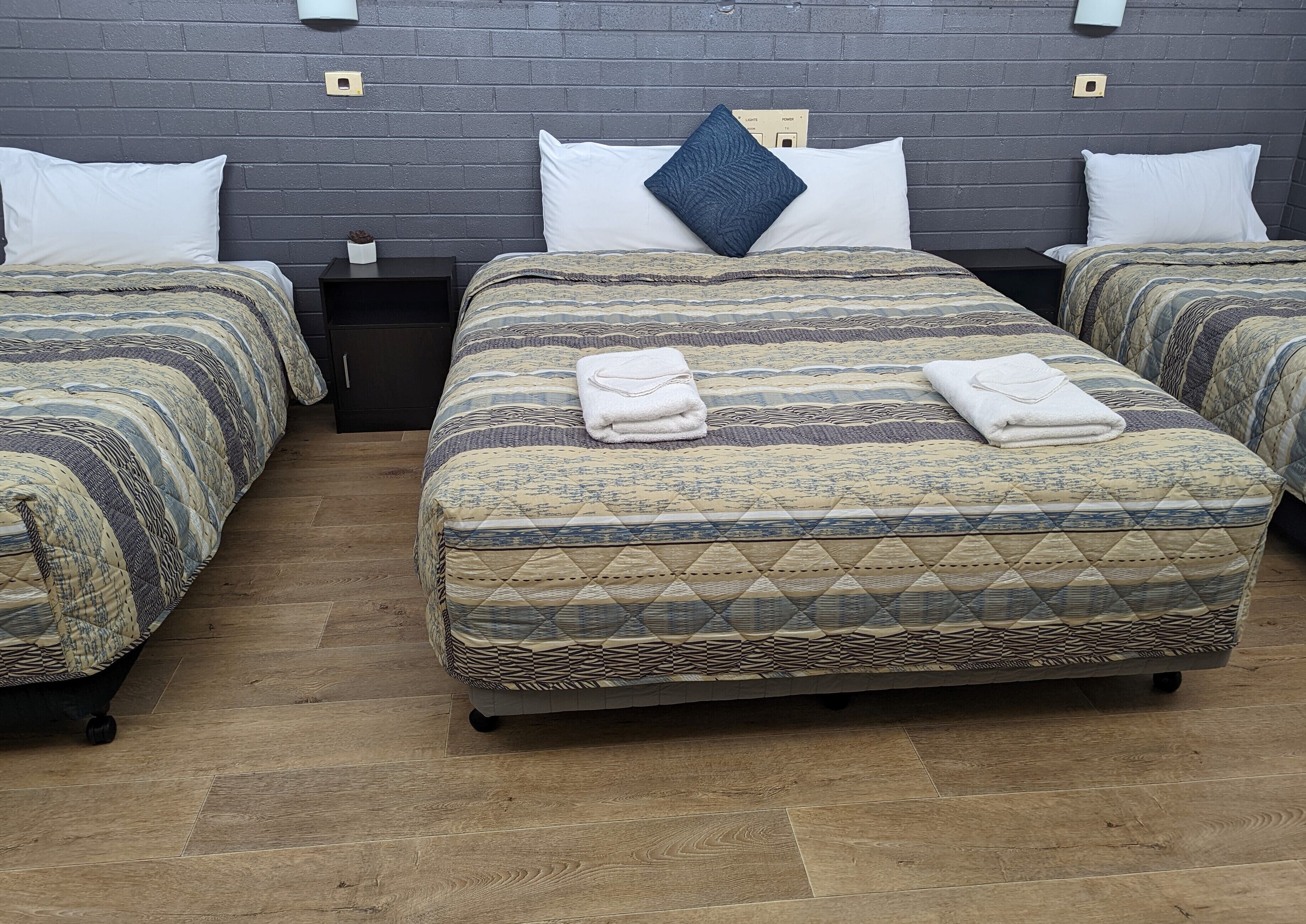 A hotel room with two beds, each with patterned bedding and a pillow. There are two nightstands with a lamp and a power outlet. The room has a wooden floor and a dark brick wall.