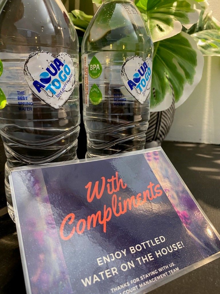 Two bottles of Aquatoo water with labels and a sign that reads "With Compliments ENJOY BOTTLED WATER ON THE HOUSE! THANKS FOR STAYING WITH US. COURT MANAGEMENT TEAM".
