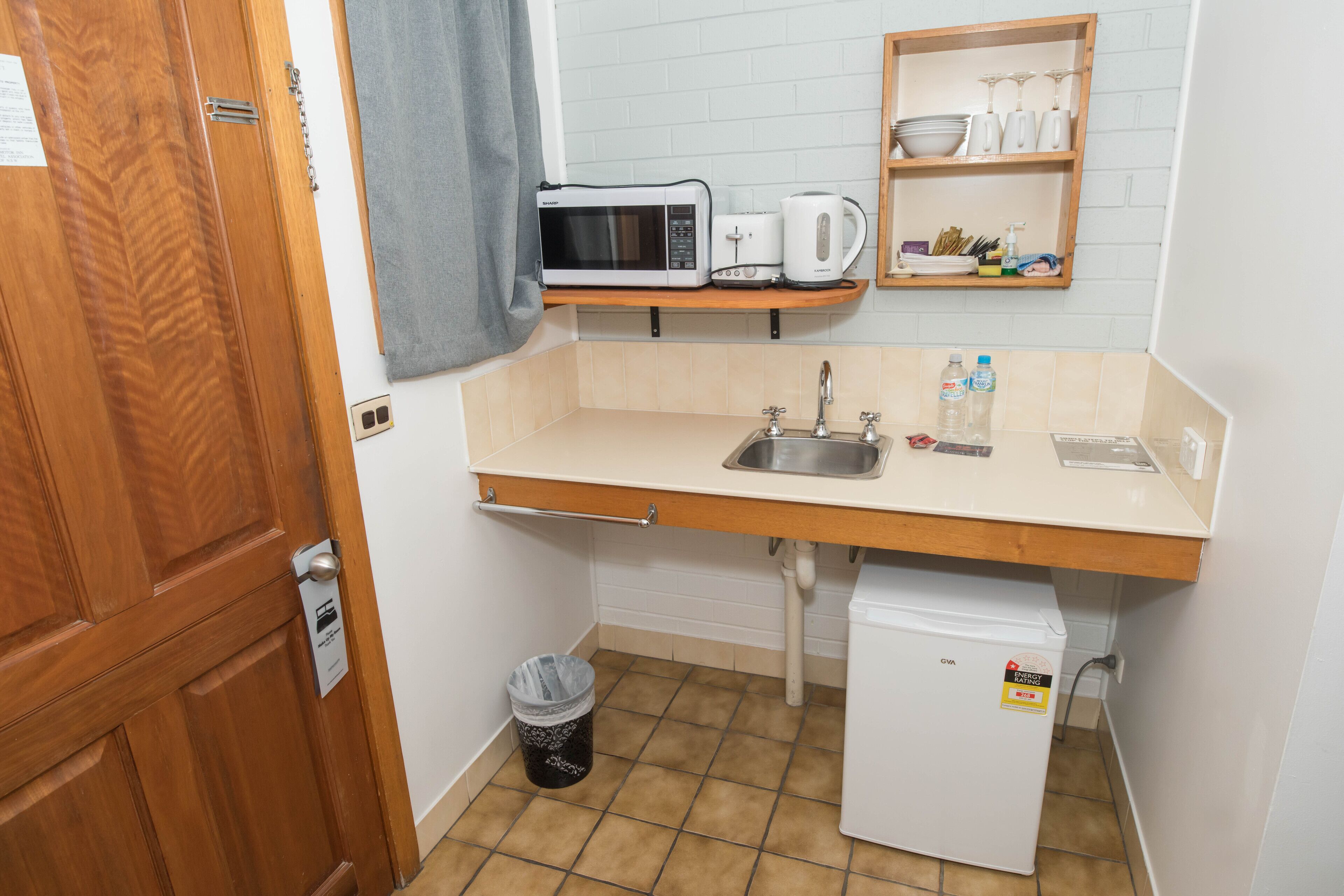 A compact kitchenette with a microwave, toaster, and coffee maker on a shelf, a sink with a faucet, a small refrigerator, and a trash bin.