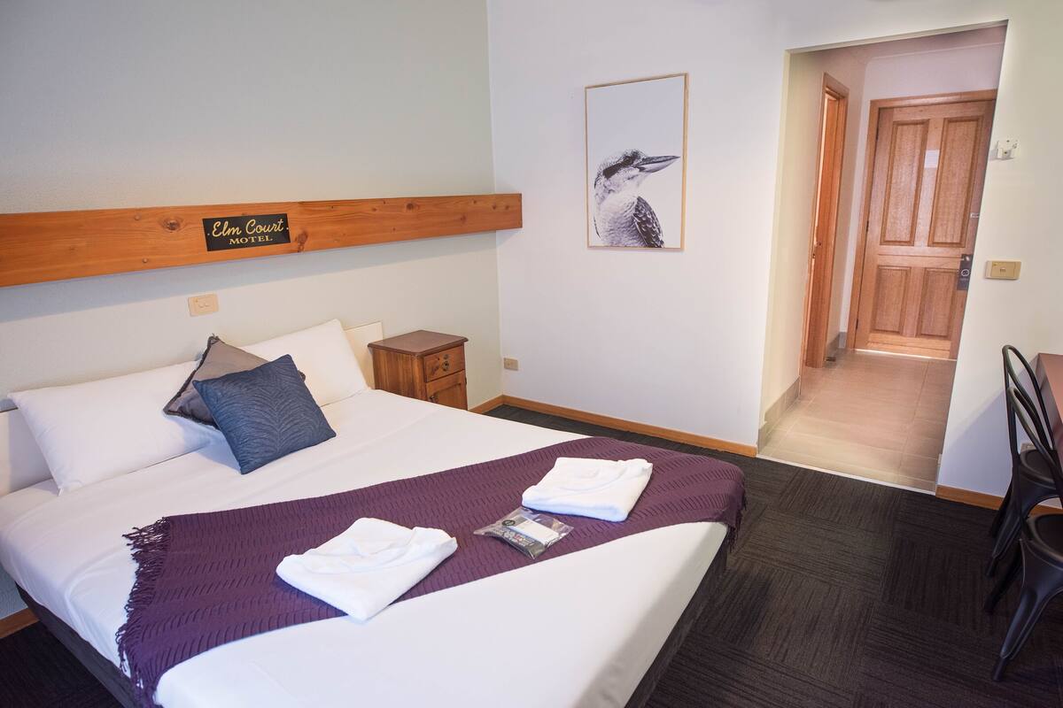 A hotel room with a bed, a wooden headboard, a pillow, a blanket, a framed picture, a nightstand, and a door leading to another room.
