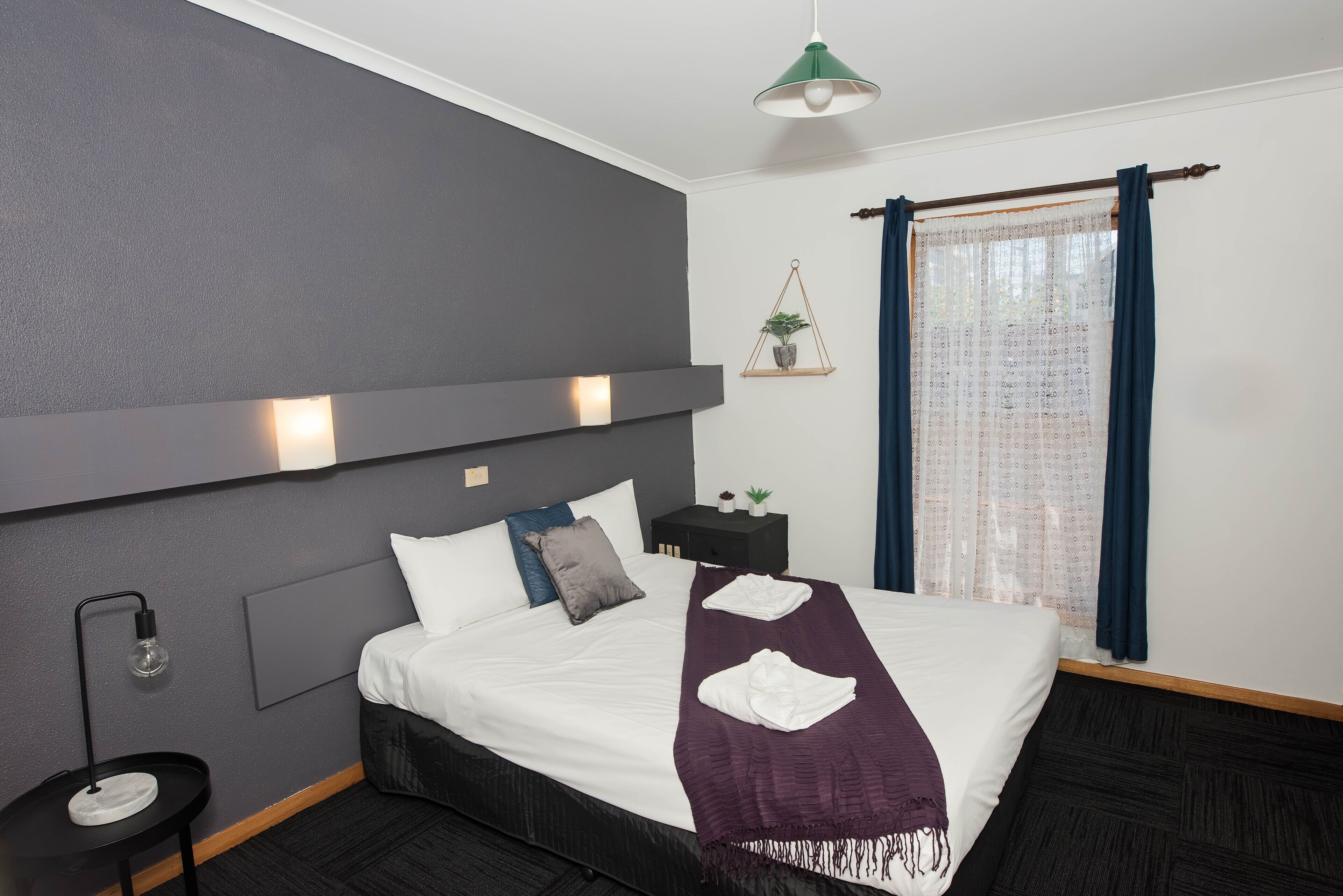 Una habitación de hotel moderna con una cama grande, una mesita de noche con lámpara, un pequeño tocador, un estante en la pared con una planta y una ventana con cortinas.