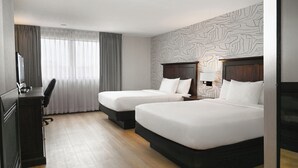 Ein Hotelzimmer mit zwei Betten, einem Schreibtisch mit Stuhl, einem Fernseher und einem Fenster mit Vorhängen.