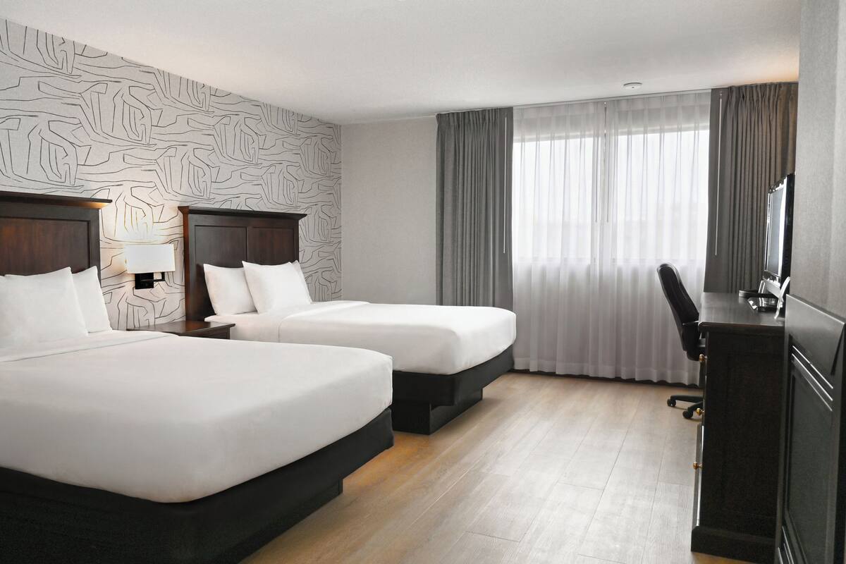 A hotel room with two beds, a desk, and a large window with curtains.