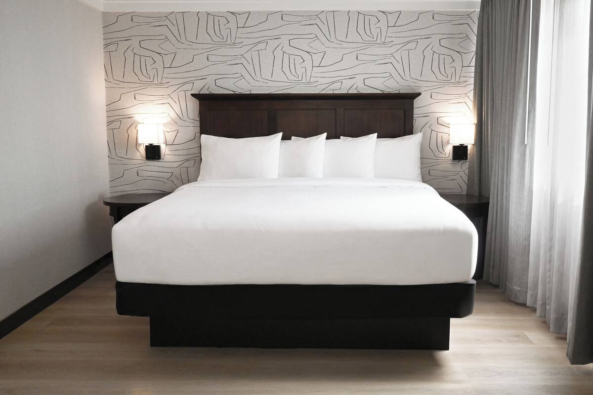 A hotel room with a large bed, two bedside tables with lamps, a dark headboard, and a patterned wall.
