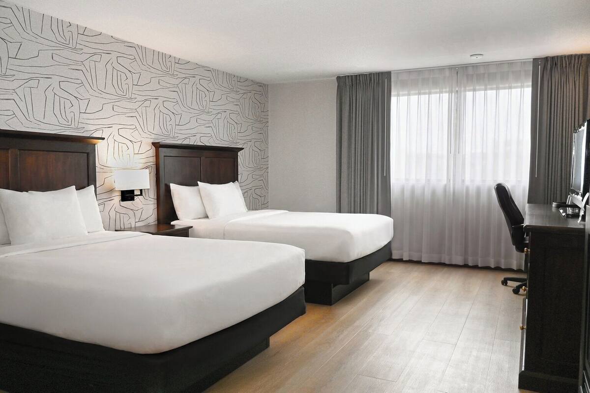 A hotel room with two beds, a desk, and a large window with curtains.
