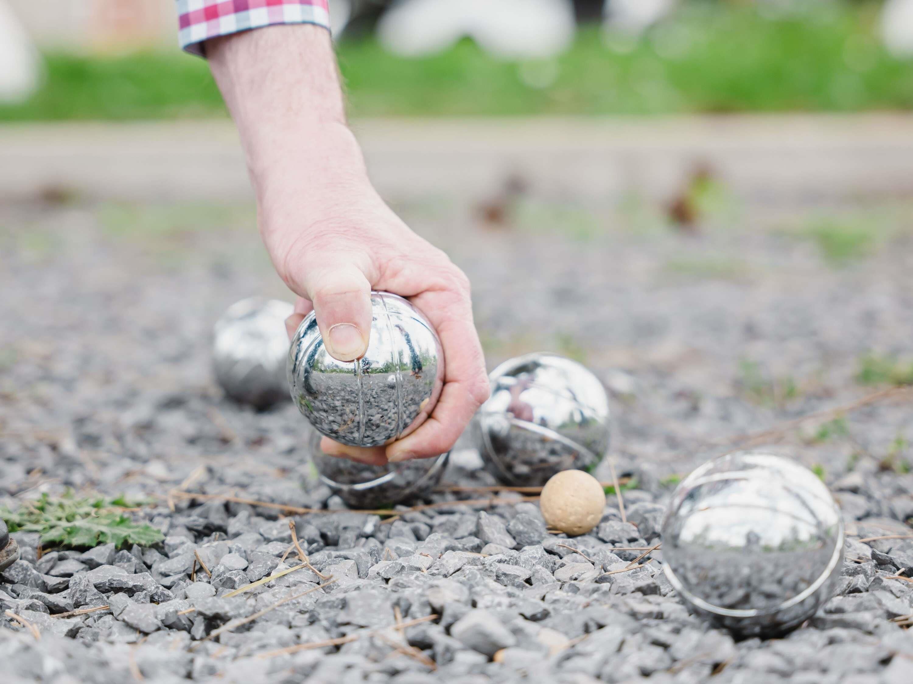 A hand holding a metallic ball on a gravel surface with other balls scattered around.
