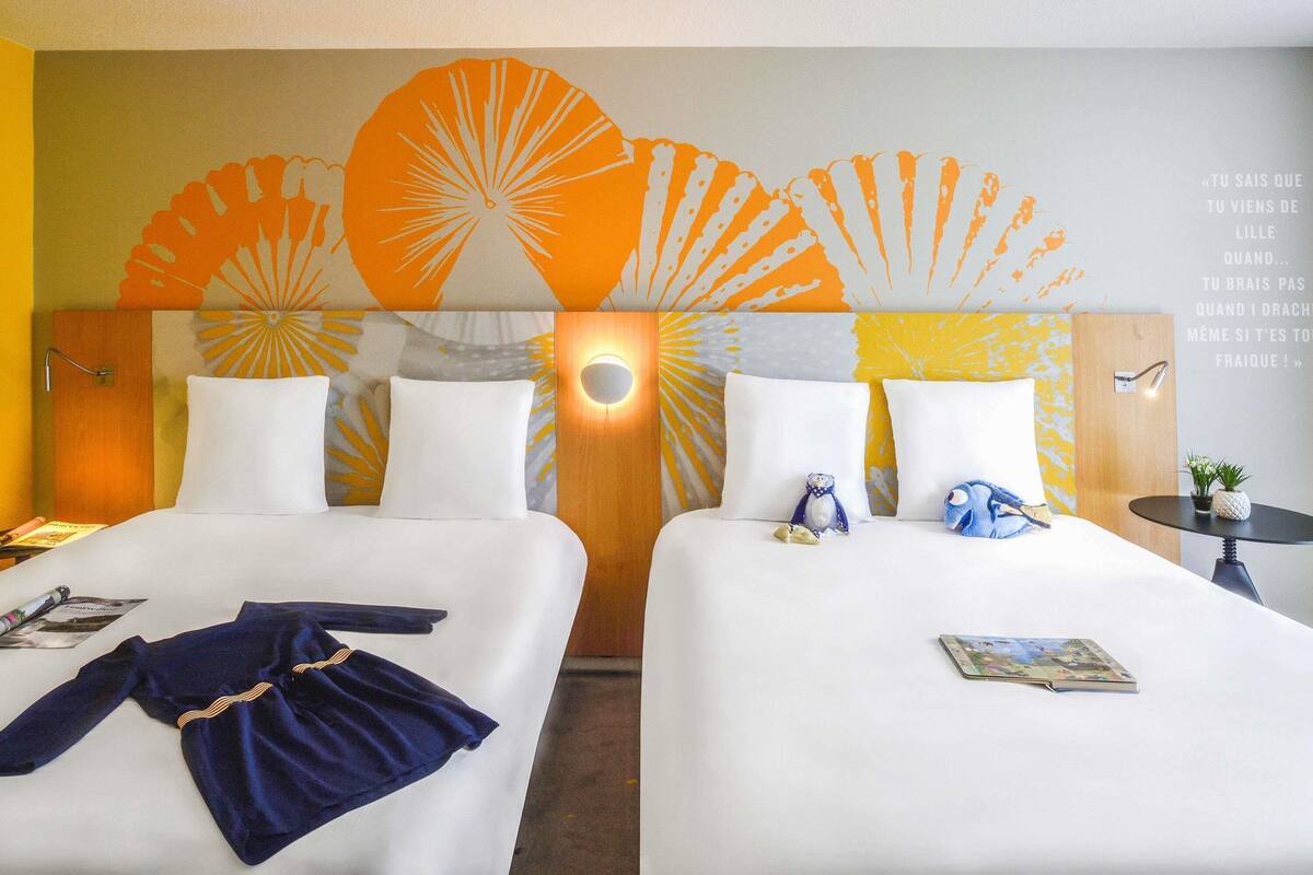 Une chambre d’hôtel avec deux lits, une tête de lit en bois ornée d’un motif en forme de soleil, un lampadaire fixé au mur et une petite table avec une plante.
