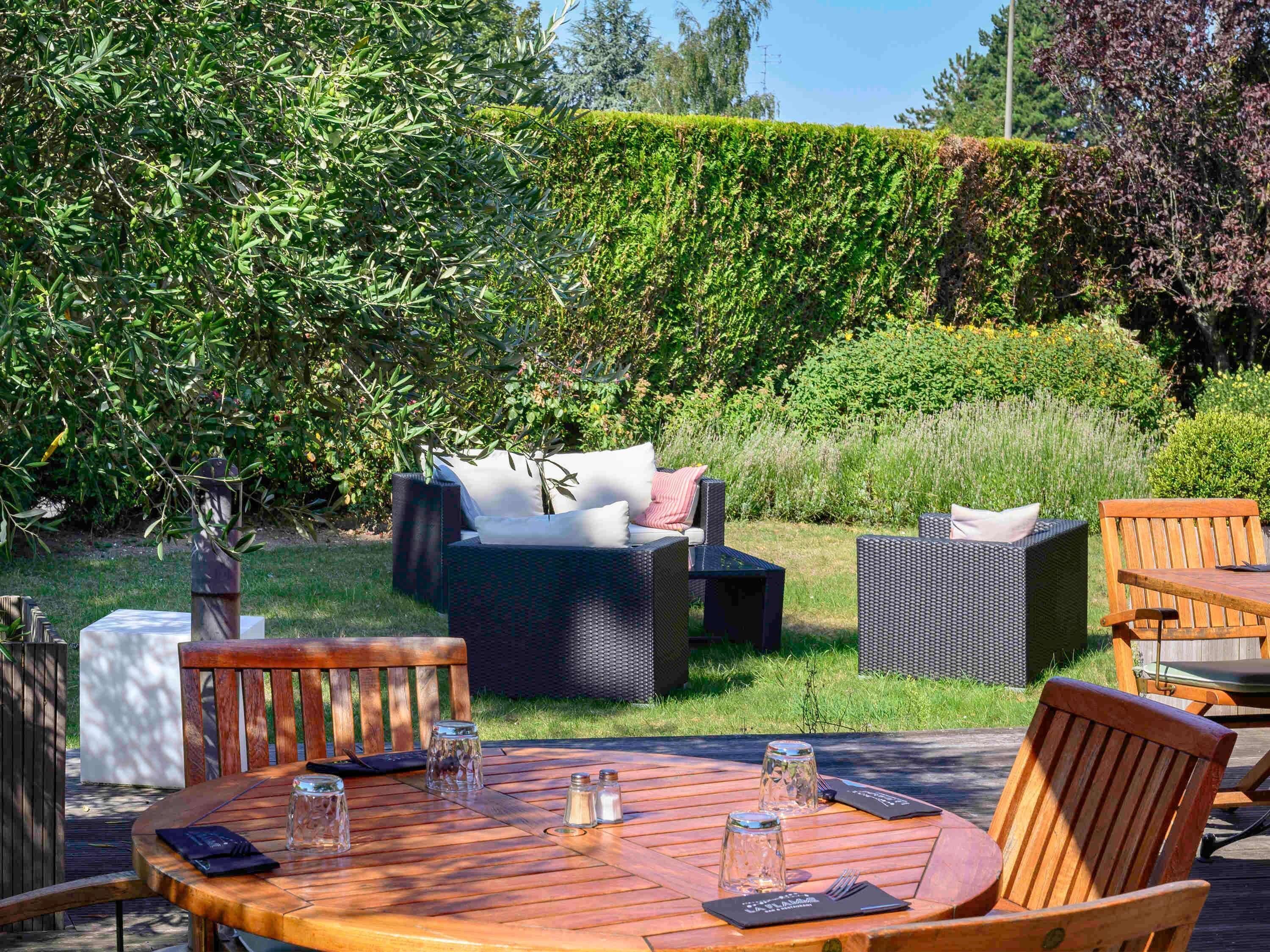 An outdoor patio with a round wooden table set for dining, surrounded by wicker furniture and a hedge in the background.