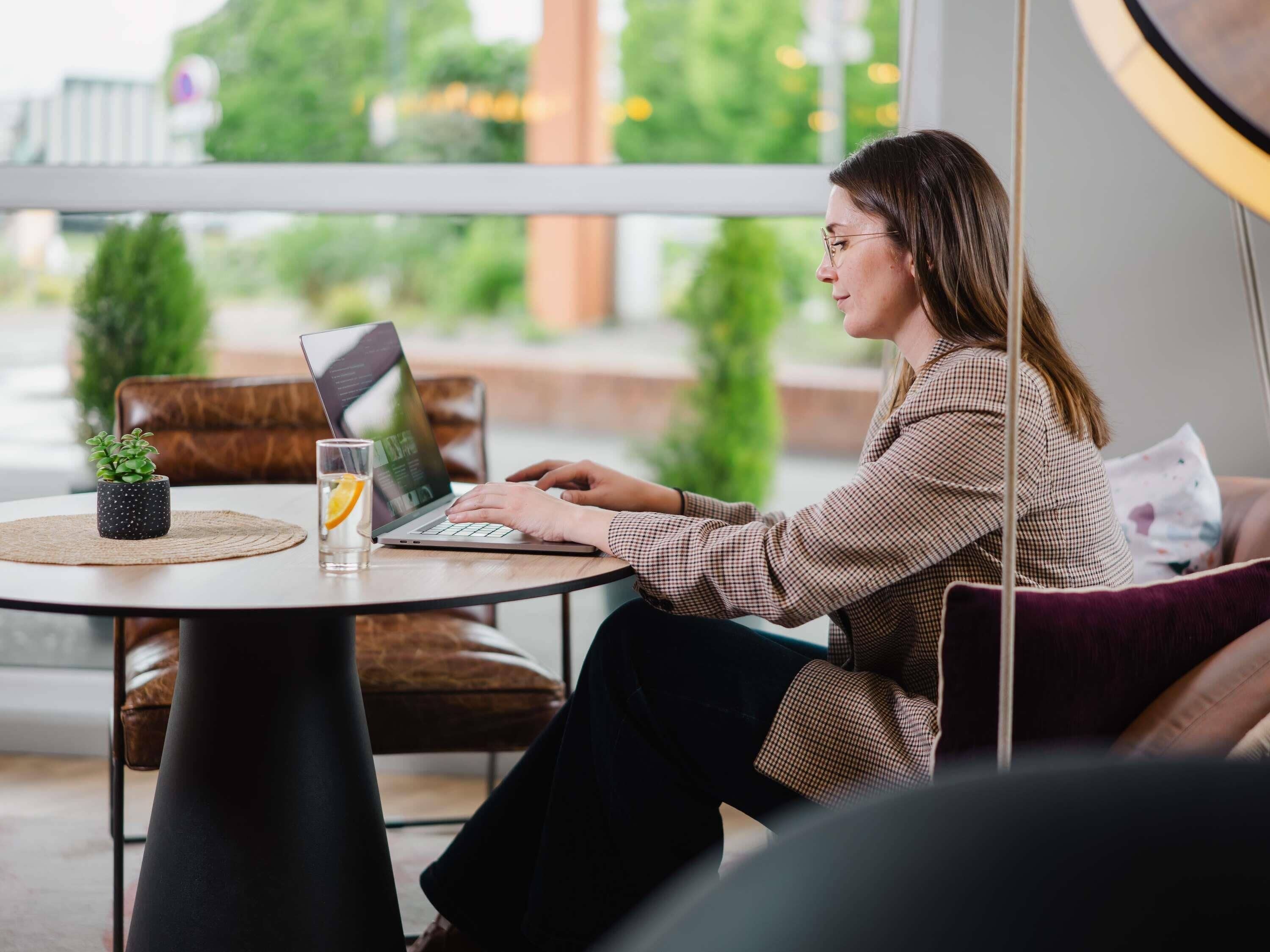 A person sitting at a round table with a laptop, a small potted plant, and a glass of water.