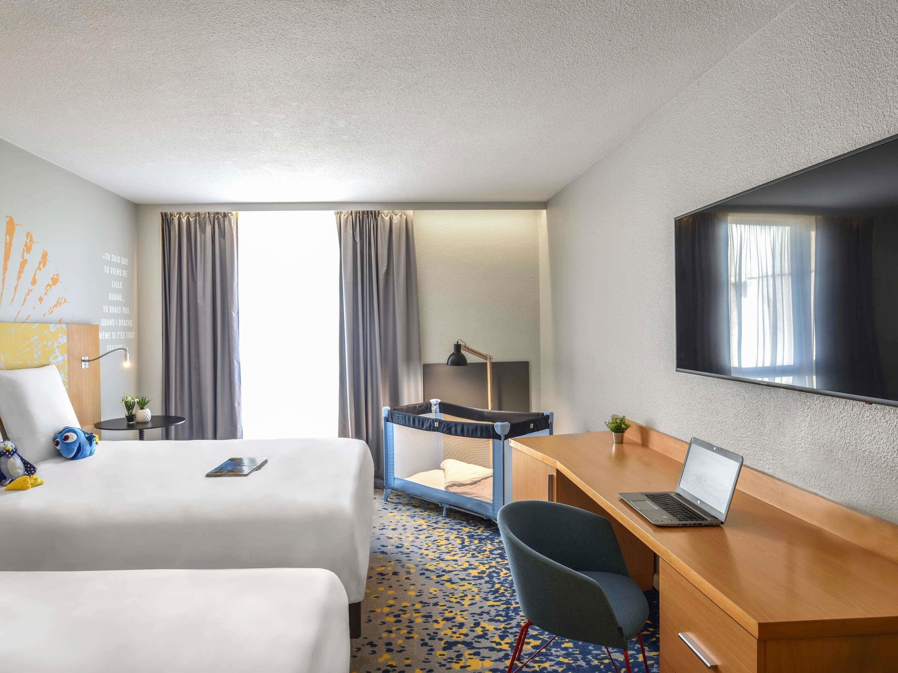 A hotel room with two beds, a desk with a laptop, a TV, and a window with curtains.