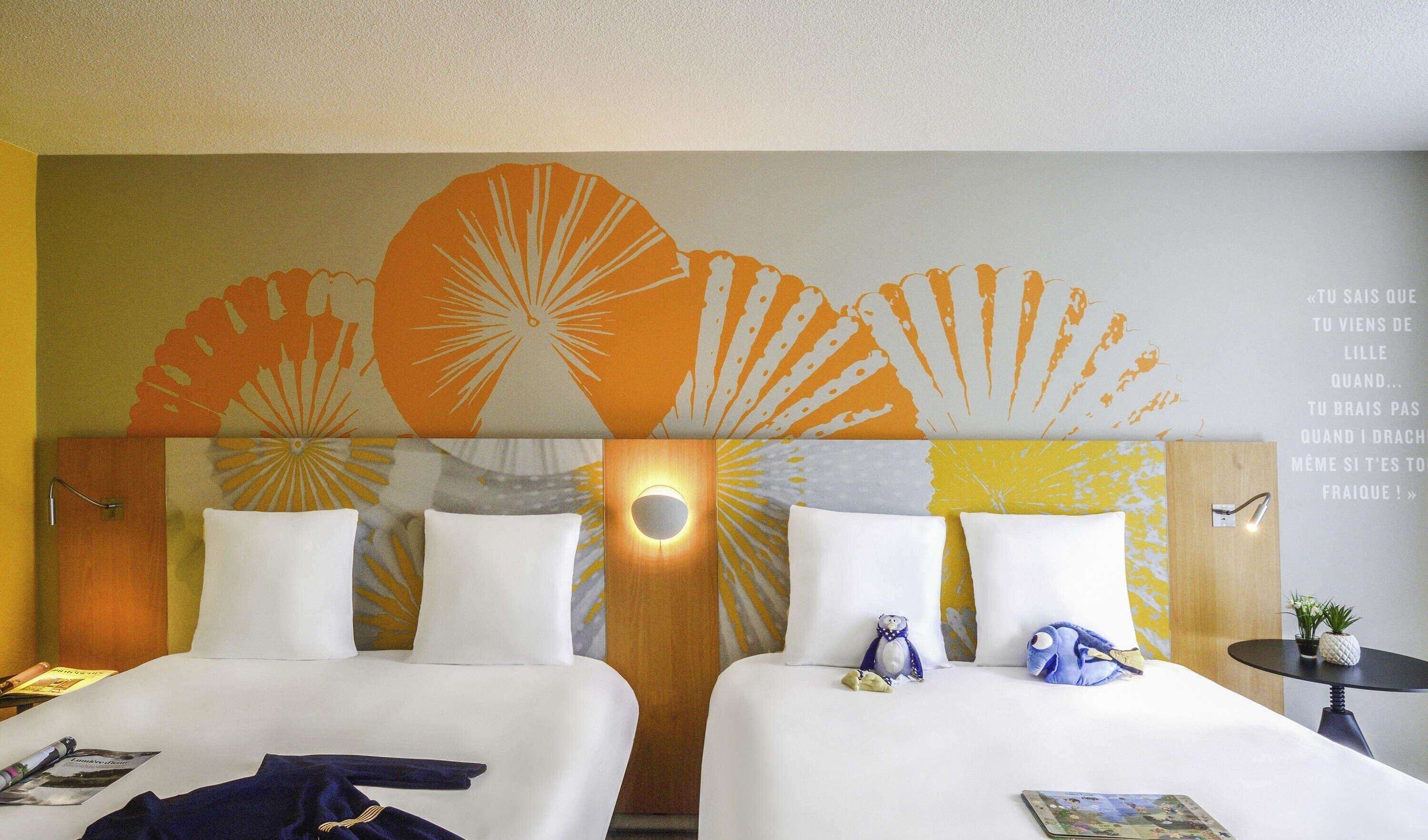 Habitación de hotel con dos camas, cabecera de madera con diseño de sol radiante, una mesita con una planta y una pared con una cita en francés.