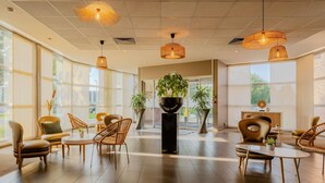 A spacious lobby with large windows, wicker chairs, a central plant, and hanging wicker lamps. - Mercure Lille Aeroport (Lesquin)