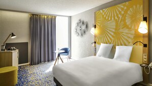 A hotel room with a bed, bedside table, lamp, chair, and a wall with a floral pattern. - Mercure Lille Aeroport (Lesquin)