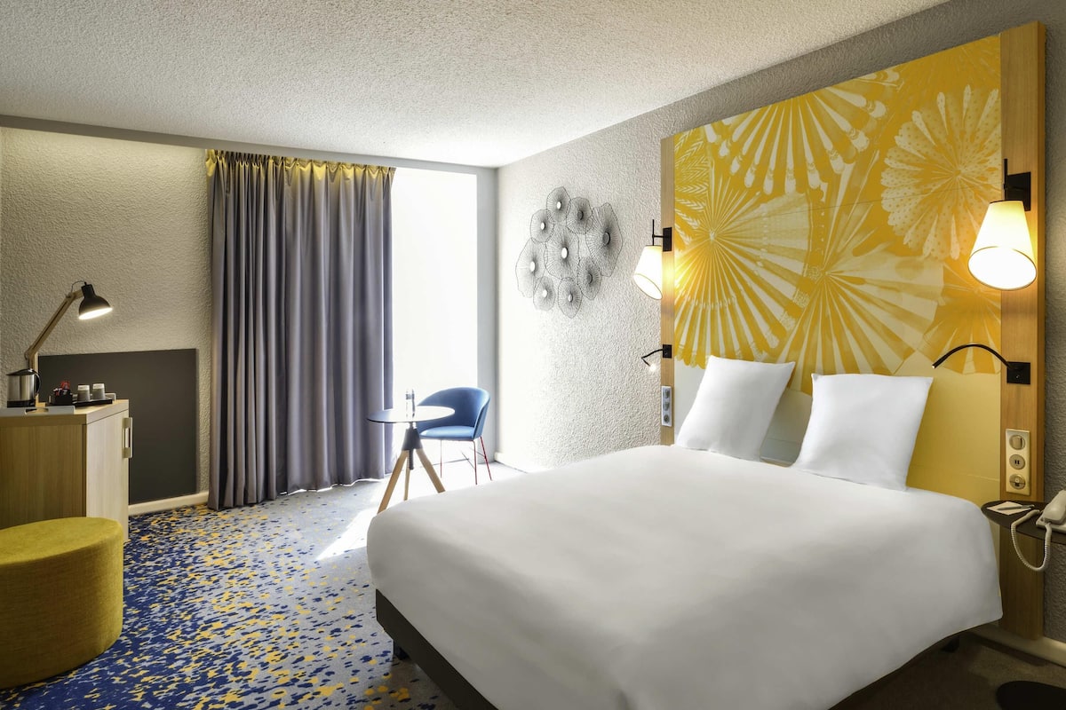 Une chambre d’hôtel comprenant un lit, une table de chevet, une lampe, une chaise et un mur orné d’un motif floral.