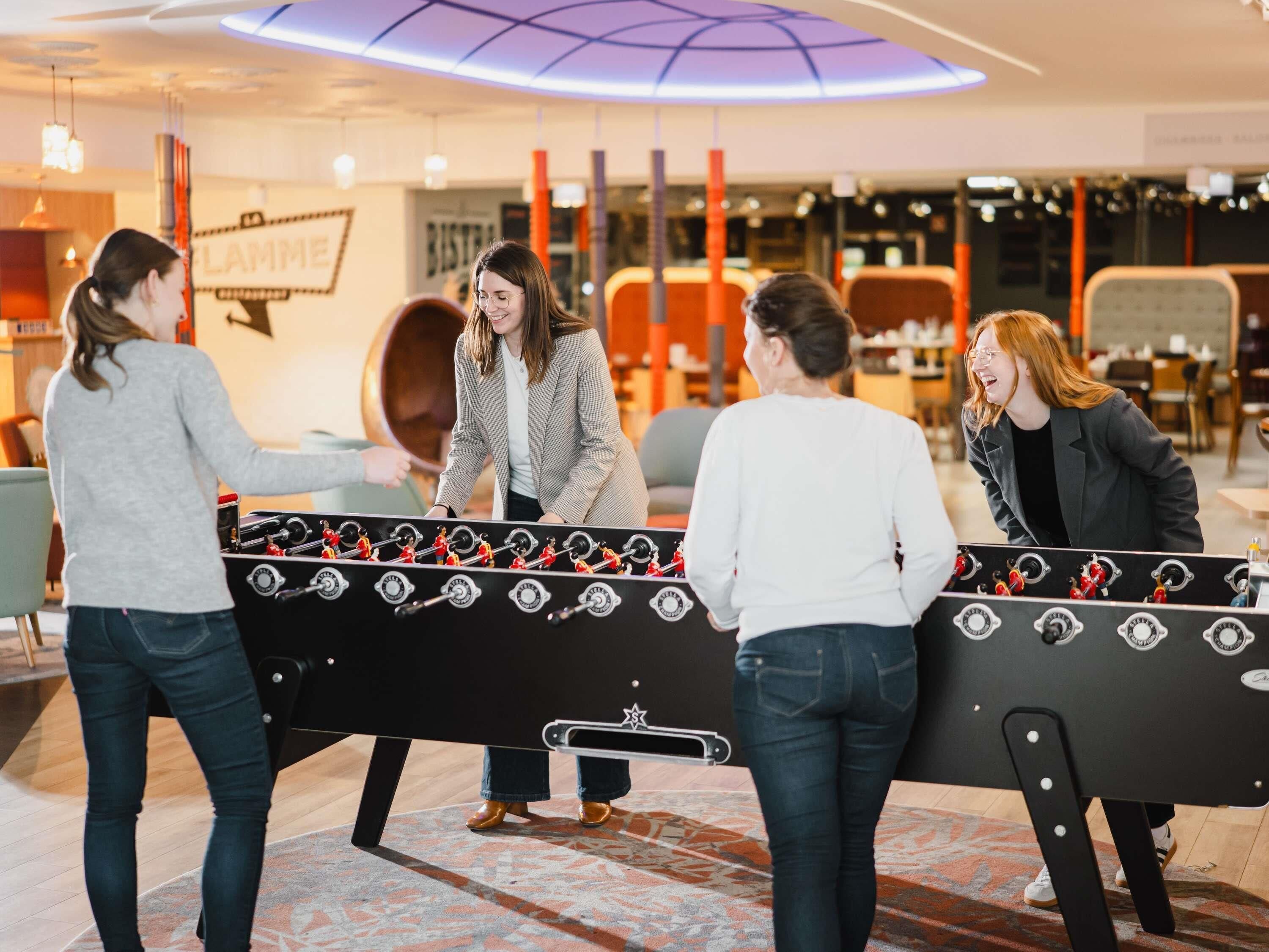 A group of people playing foosball in a modern indoor setting with a visible restaurant sign in the background.