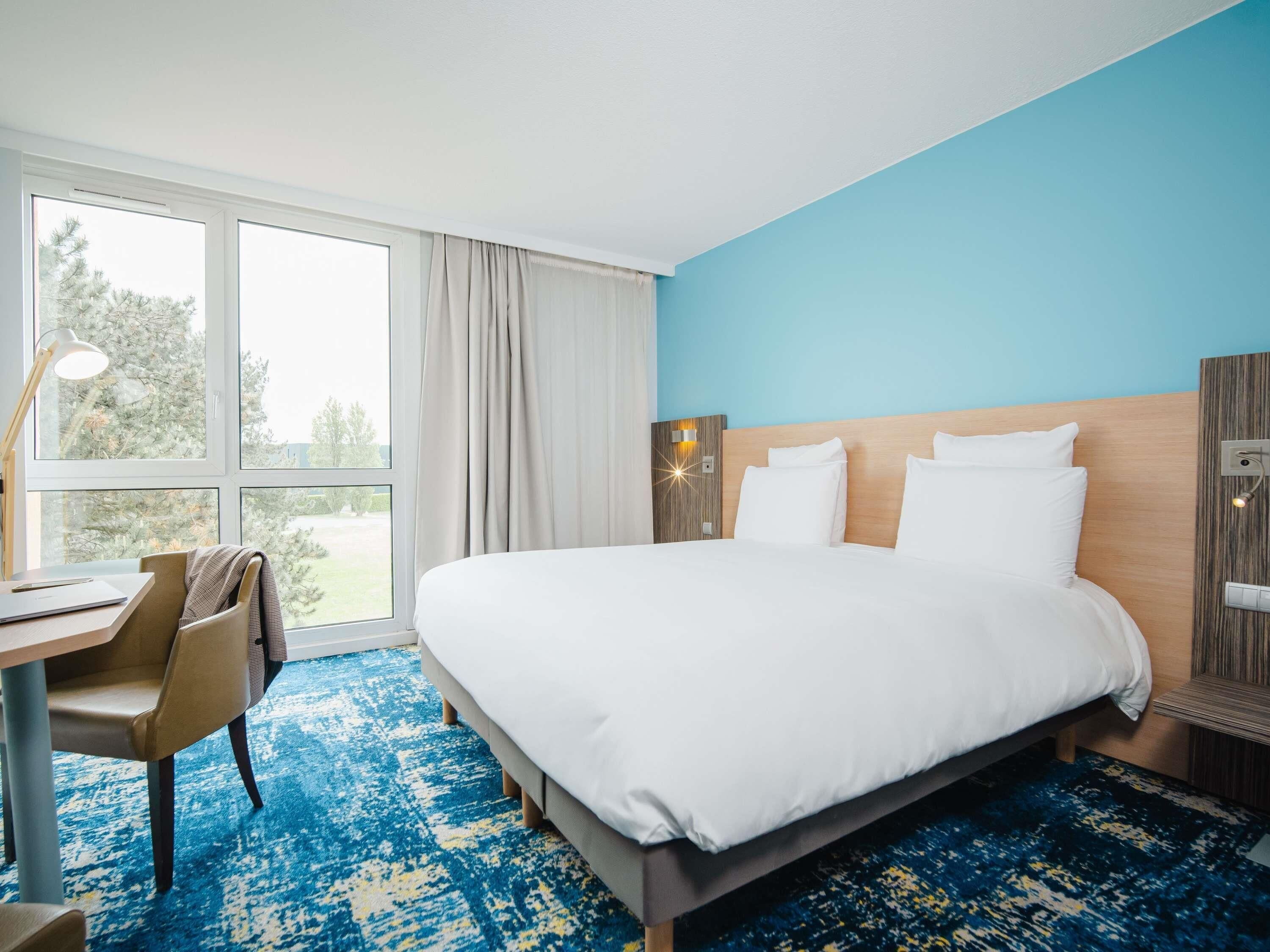 A hotel room with a large bed, a desk with a chair, a lamp, and a view of trees outside.