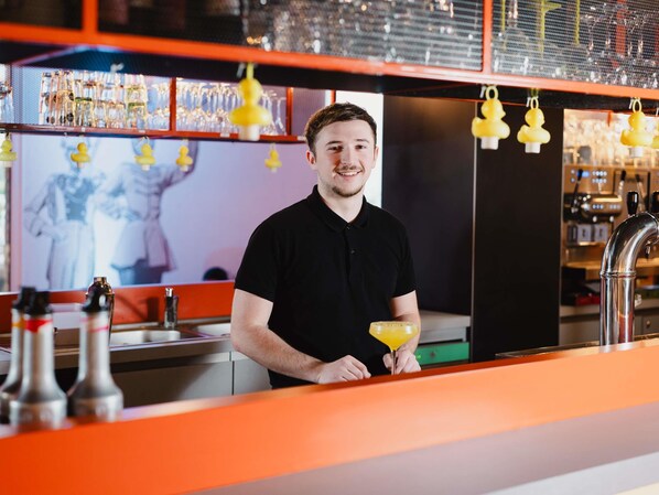 A bar area with a bartender holding a cocktail shaker, shelves with bottles, and a large screen displaying a person.