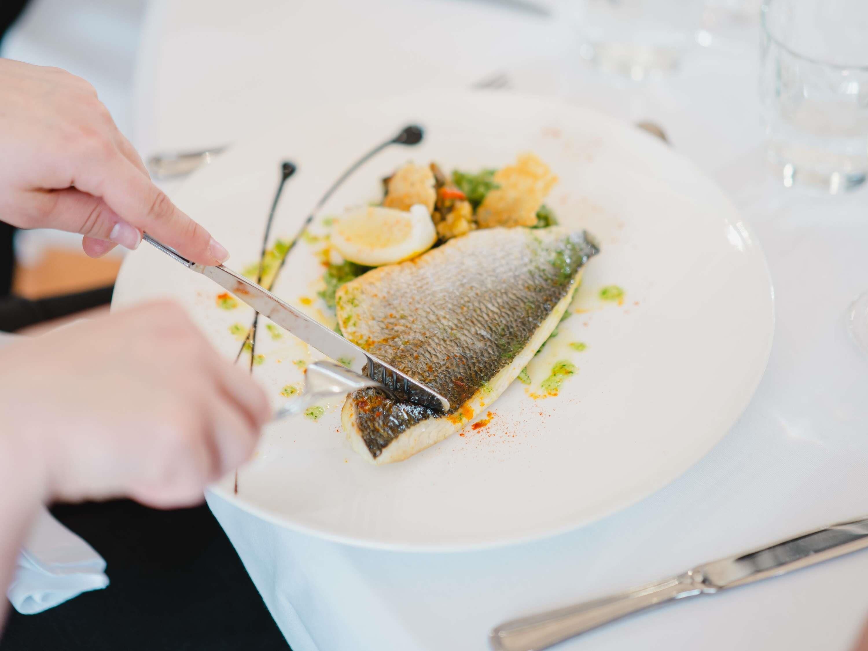 Una persona cortando un pescado en un plato blanco con un tenedor y un cuchillo.