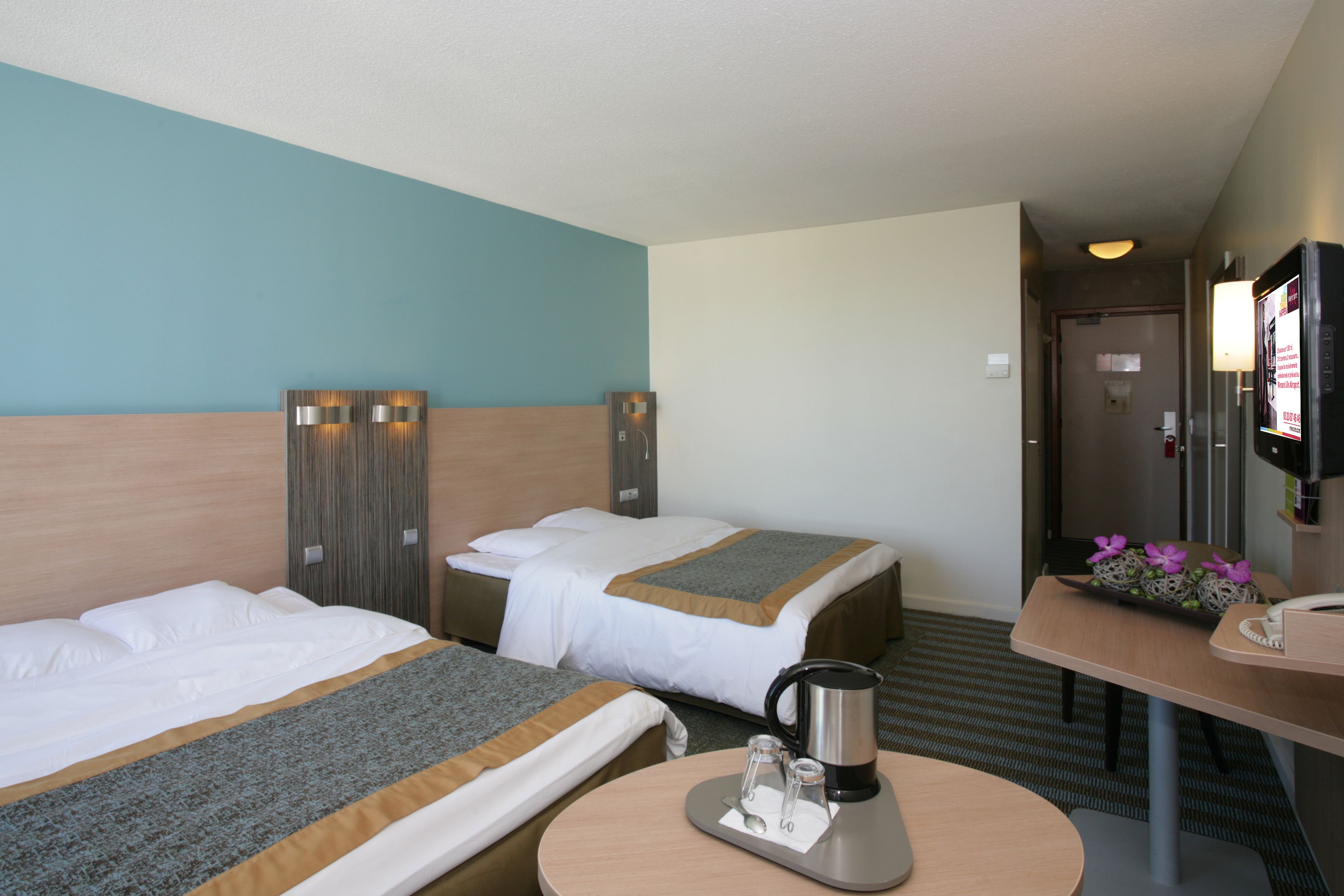 Quarto de hotel com duas camas, uma escrivaninha com cafeteira e televisão na parede.