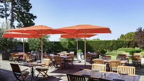 An outdoor patio with wooden tables and chairs, orange umbrellas, and a wooden deck. - Mercure Lille Aeroport (Lesquin)