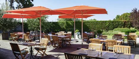 An outdoor patio with wooden tables and chairs, orange umbrellas, and a wooden deck.