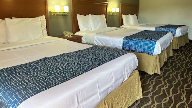 Ein Hotelzimmer mit drei Betten, jedes mit weißer Bettwäsche und einem blau gemusterten Überwurf. An beiden Seiten der Betten befinden sich Wandlampen, und auf dem Nachttisch steht ein Telefon.
