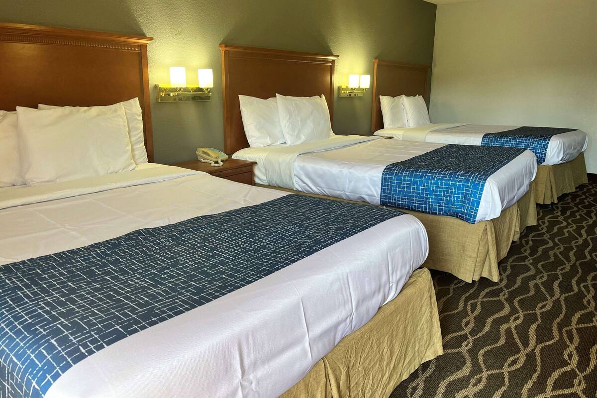 Ein Hotelzimmer mit drei Betten, jedes mit weißer Bettwäsche und einem blau gemusterten Überwurf. An beiden Seiten der Betten befinden sich Wandlampen, und auf dem Nachttisch steht ein Telefon.