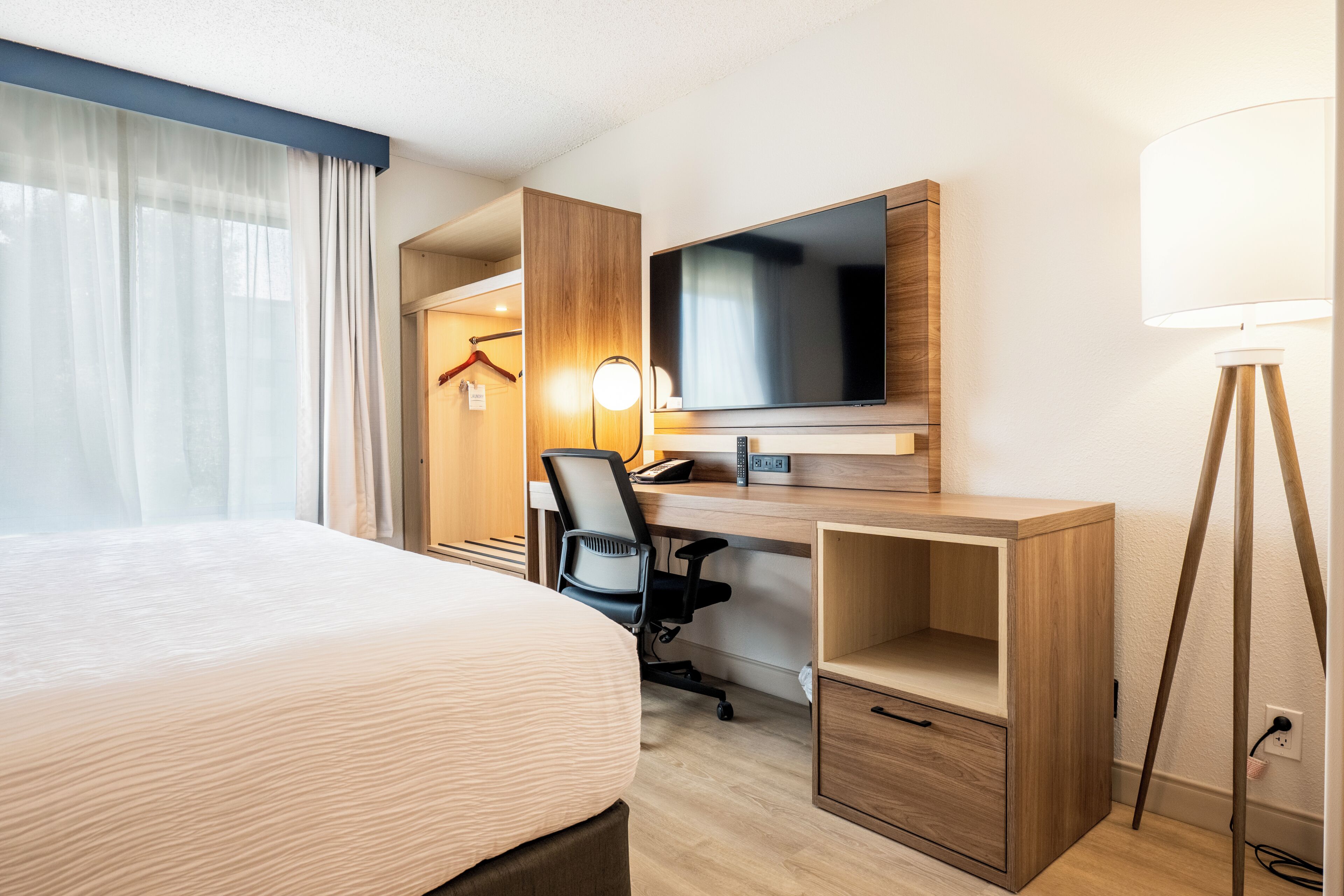 A hotel room with a bed, desk, chair, TV, lamp, and wardrobe.