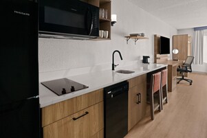 Cocina moderna con refrigerador negro, microondas, lavamanos y lavavajillas. Cuenta con encimera de madera, una silla y un televisor montado en la pared.