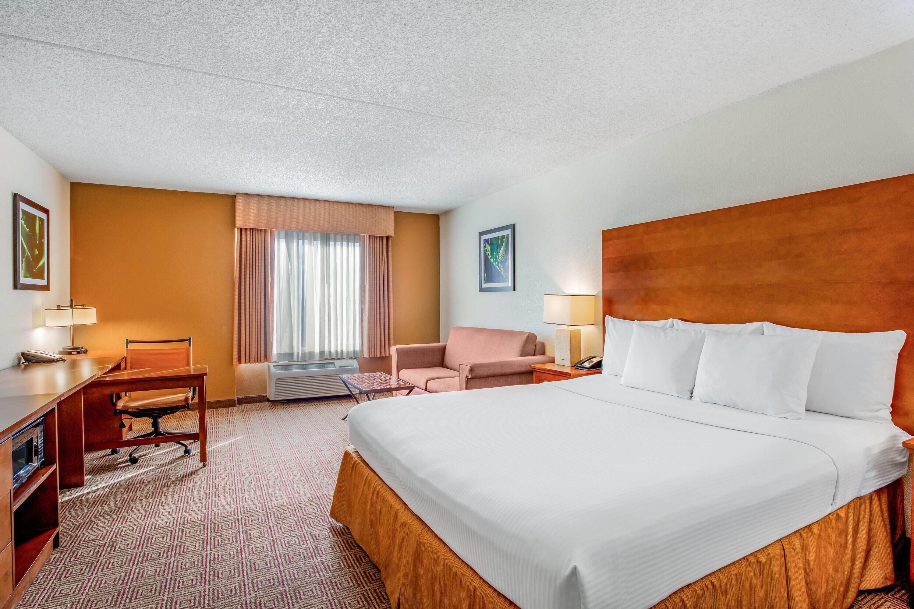 Ein Hotelzimmer mit einem großen Bett, einem Schreibtisch, einem Sessel, einem kleinen Tisch und einem Fenster mit Vorhängen.