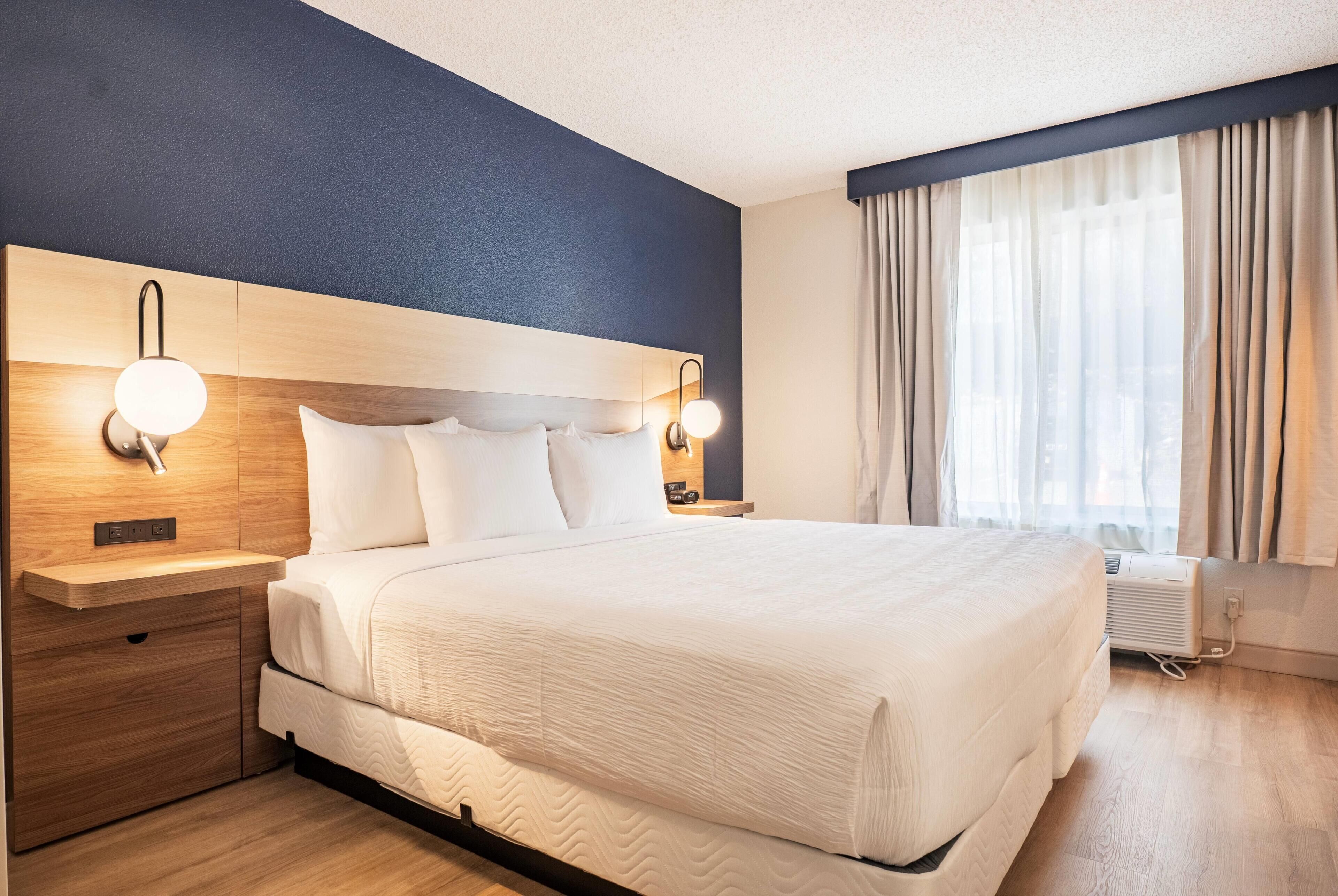 A hotel room with a large bed, wooden headboard, bedside lamps, a nightstand, and a window with curtains.