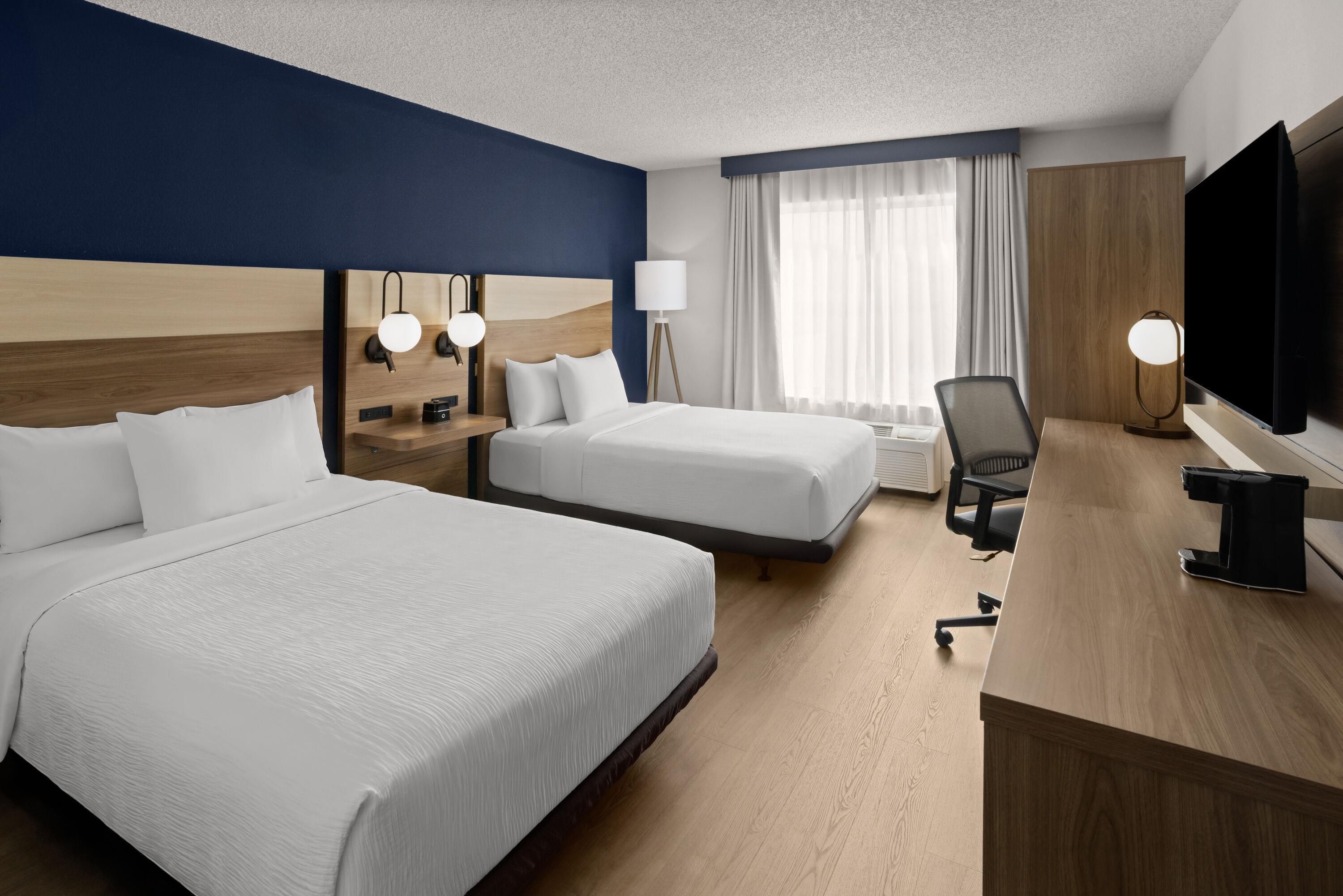 A hotel room with two beds, a desk with a chair, a TV, and a window with curtains.