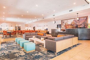 A modern hotel lobby with a seating area, a reception desk, and a kitchen in the background.