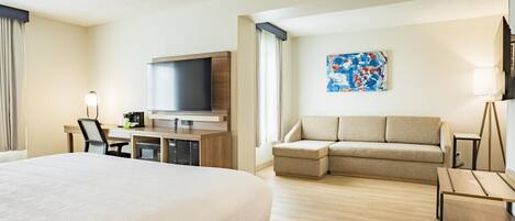 A hotel room with a large bed, a flat-screen TV, a sofa, a desk with a lamp, and a painting on the wall.