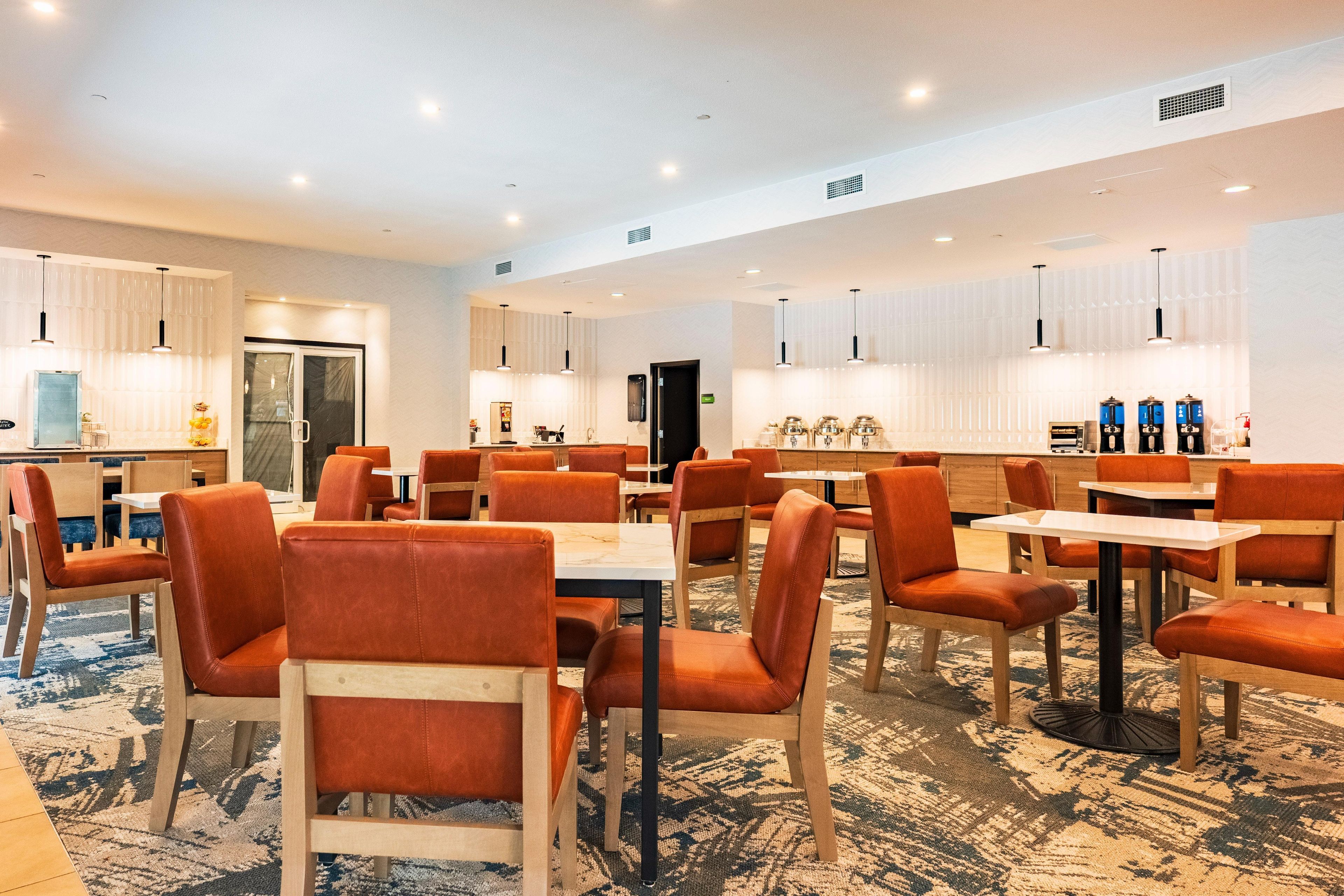 Un comedor de hotel con varias mesas y sillas, una cocina al fondo y una alfombra estampada.
