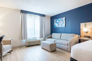 Habitación de hotel con sofá, reposapiés, mesita de noche, lámpara, aire acondicionado y un cuadro en la pared.