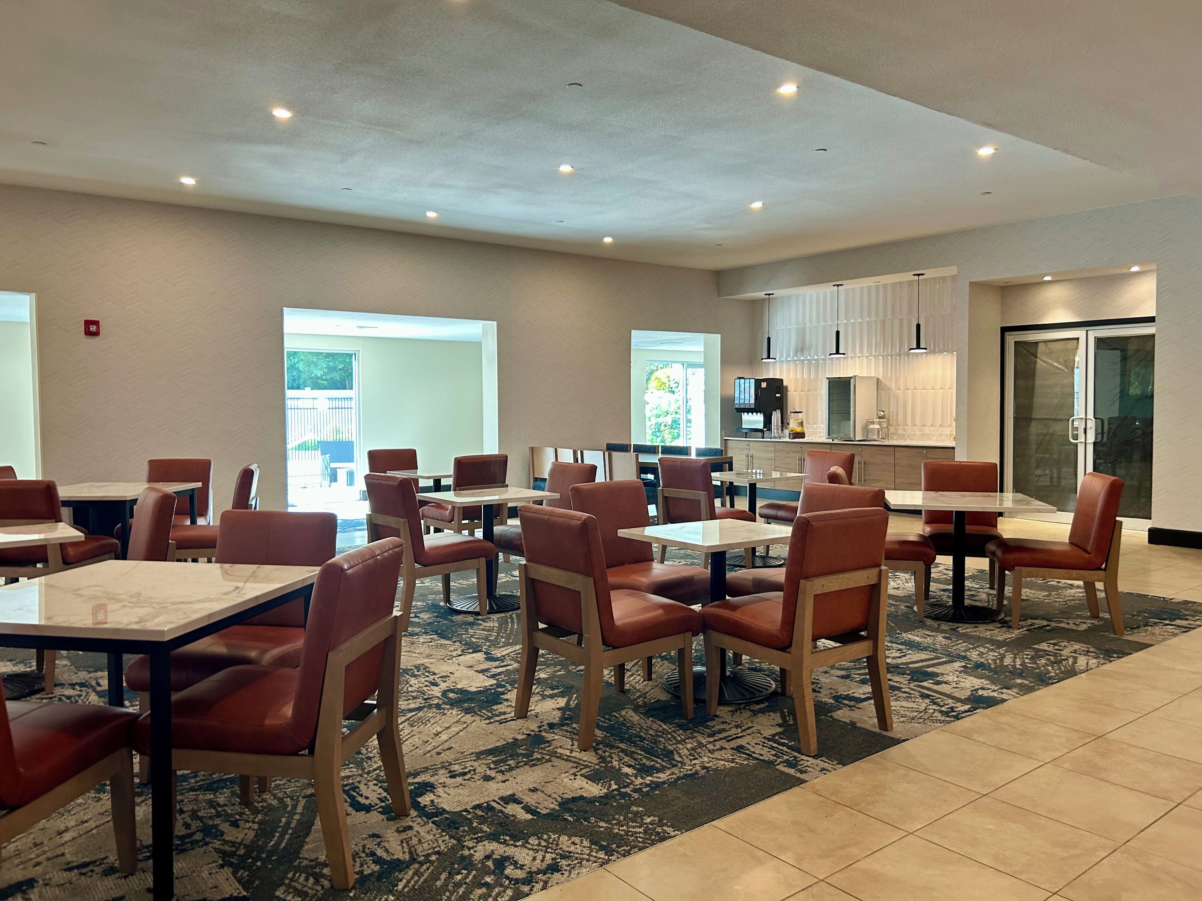 A hotel dining area with multiple tables and chairs, a kitchen area, and a view of the outdoors.
