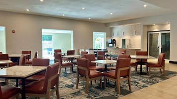 A hotel dining area with multiple tables and chairs, a kitchen area, and a view of the outdoors.