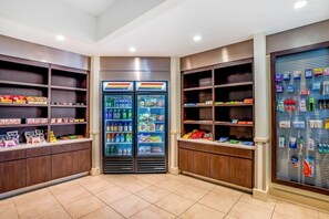 Une pièce spacieuse et bien éclairée, dotée d’un sol carrelé, et comprenant un grand réfrigérateur à double porte placé au centre, rempli de boissons et d’en-cas, et entouré d’étagères où sont disposés divers produits emballés.