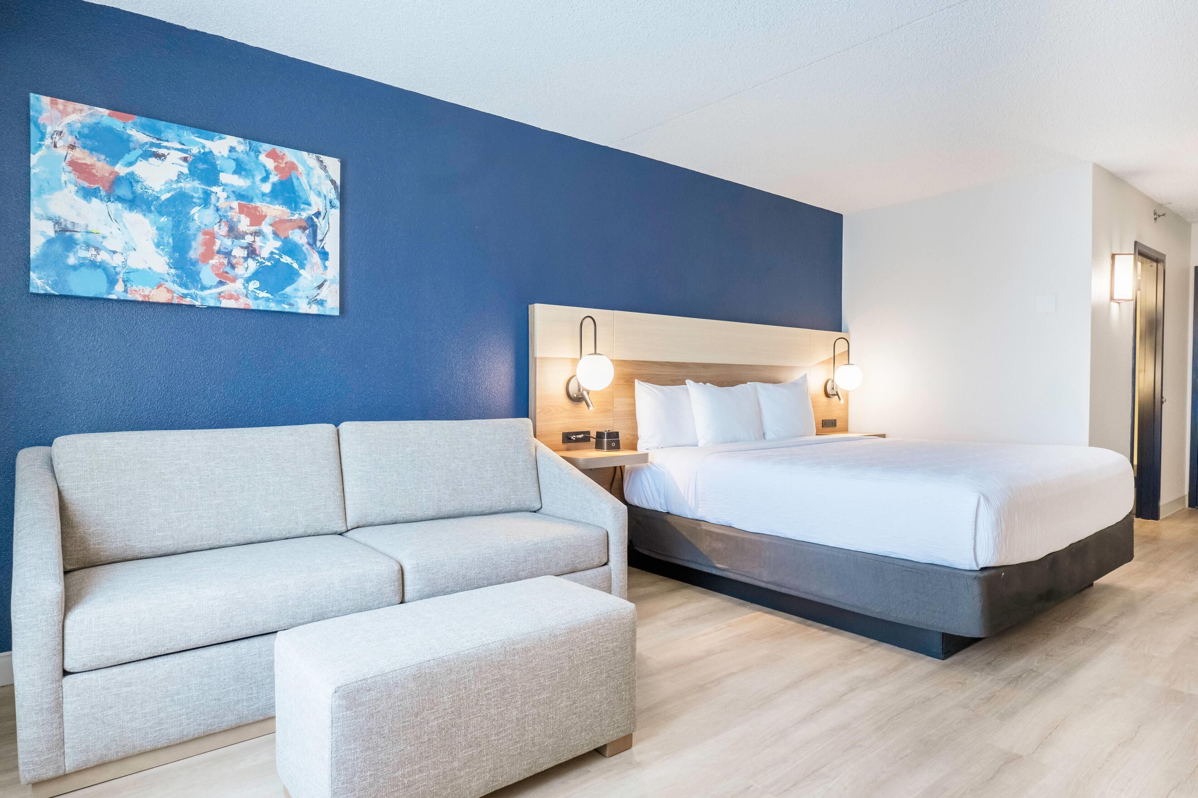A modern hotel room with a bed, a sofa, a small table, and a wall-mounted artwork.