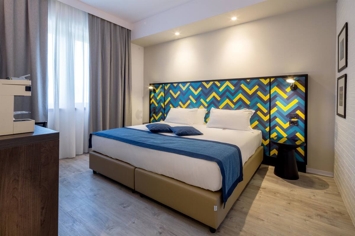 A modern hotel room with a large bed, a desk, and a view of the city.
