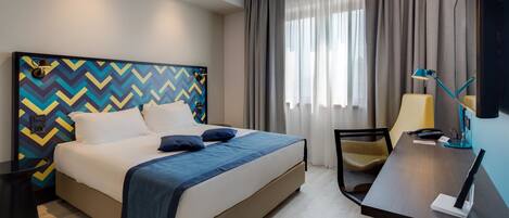 Quarto de hotel com cama, escrivaninha, cadeira e televisão.