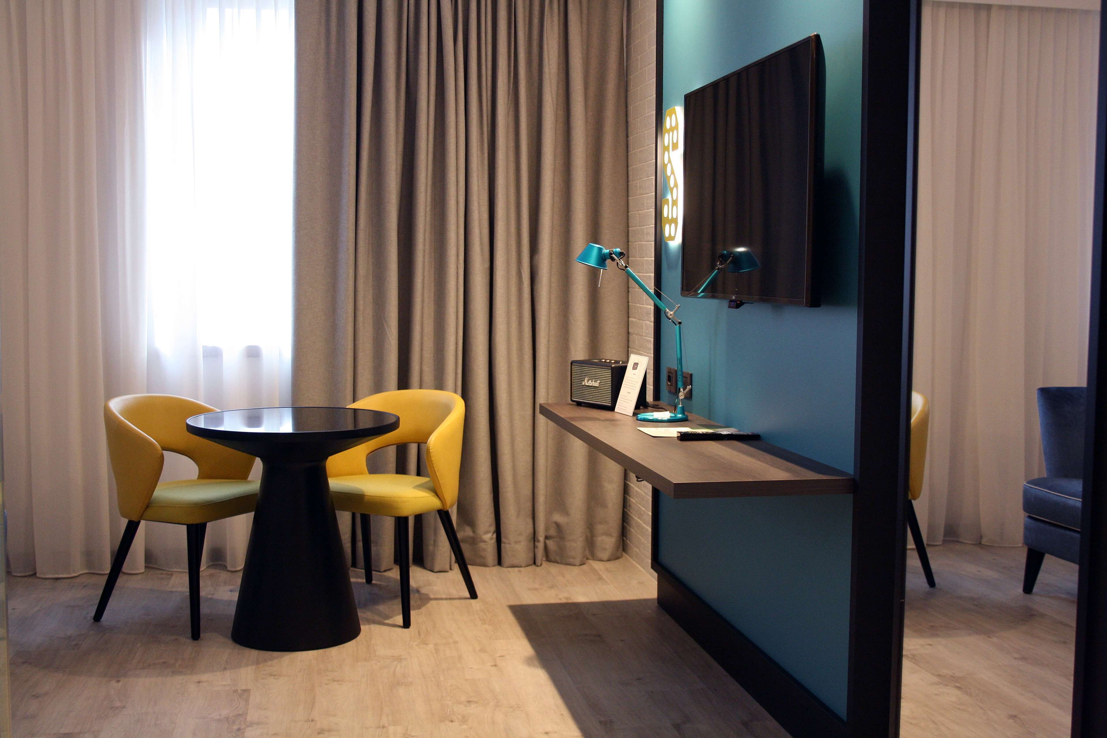 Una habitación moderna con una mesa redonda, dos sillas amarillas, una lámpara azul y un televisor de pantalla plana montado en la pared.
