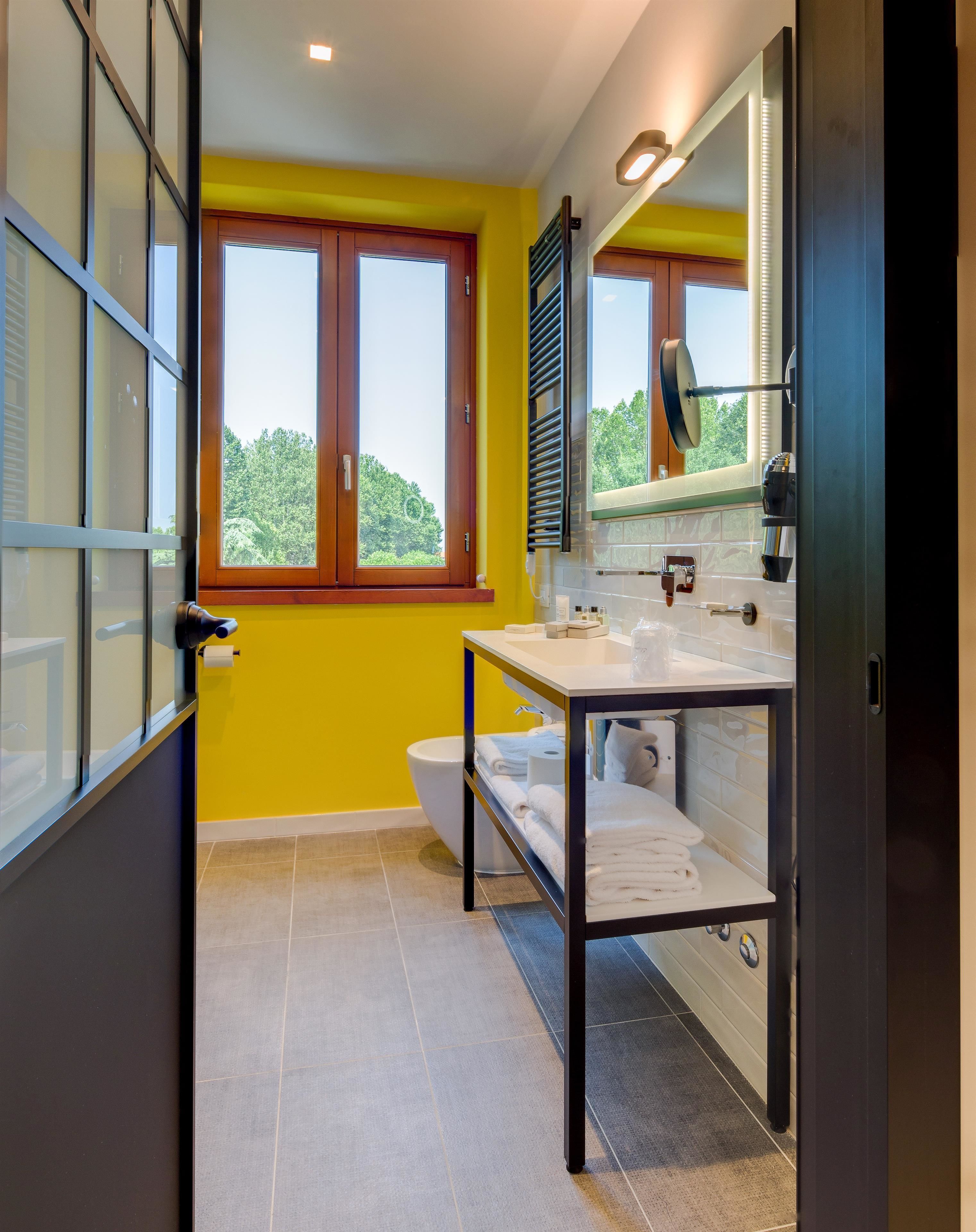 Un baño moderno con una pared amarilla, un ventanal grande, un lavamanos blanco y un espejo.