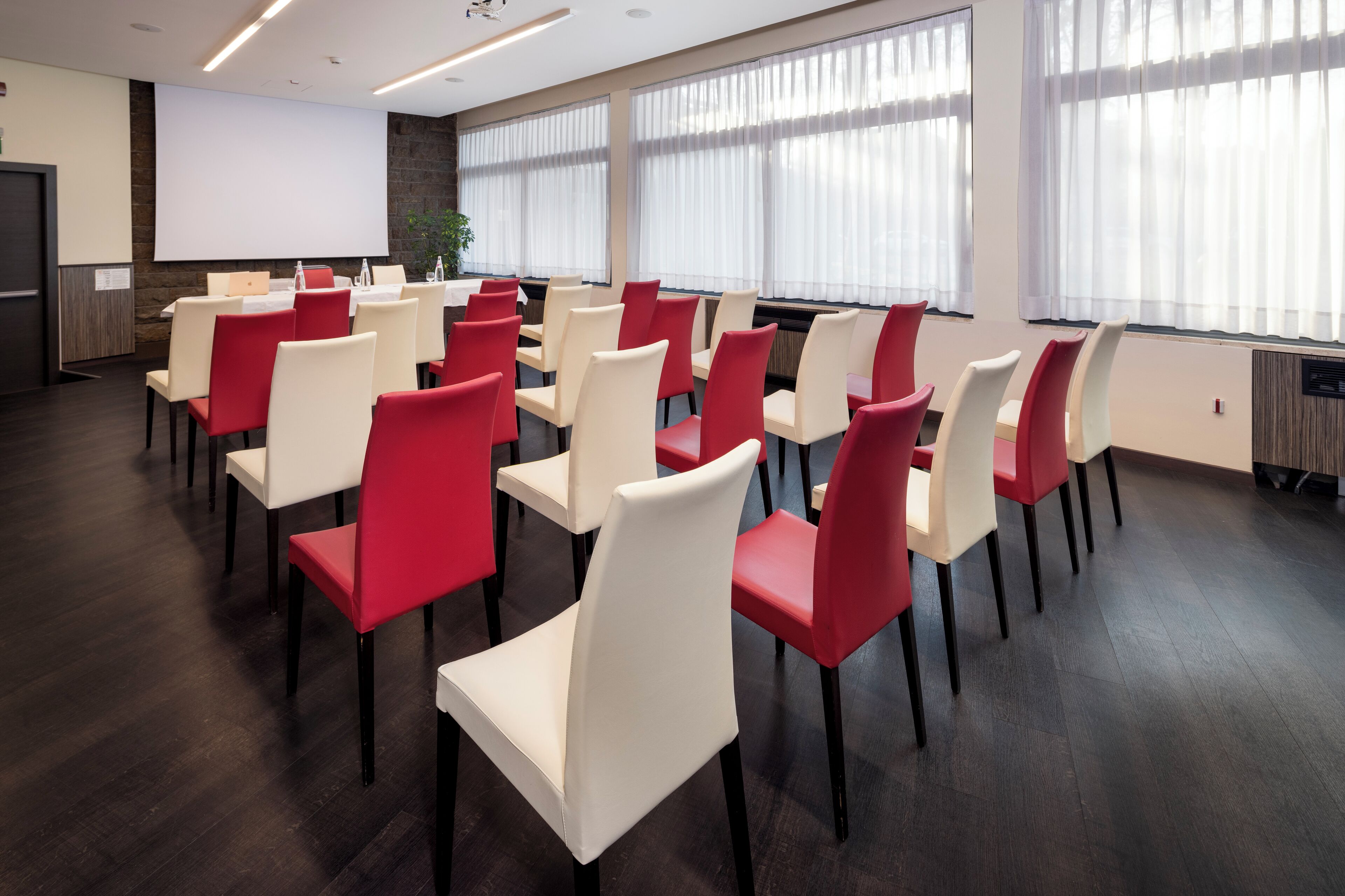 A conference room with rows of white and red chairs, a large screen, and a plant.