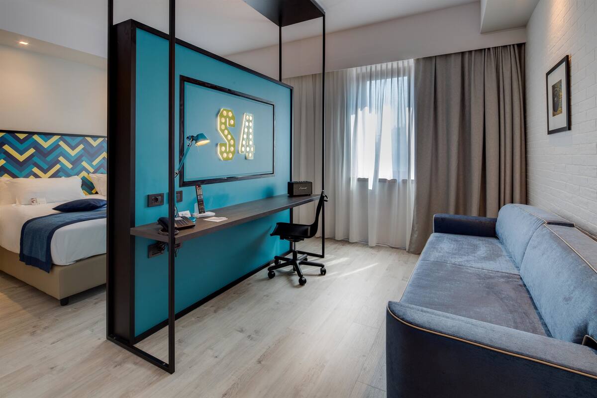 A modern hotel room with a bed, a desk with a computer, a sofa, and a large window with curtains.